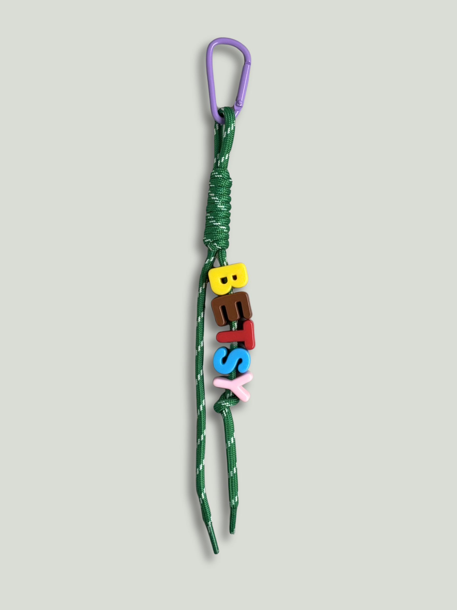 Personalised bag charm with letter beads spelling BETSY in yellow, brown, red, blue and pink, threaded on a green and white fleck knotted cord, with a purple carabiner clip on a yellow background. Made by Bugalugs in Melbourne.