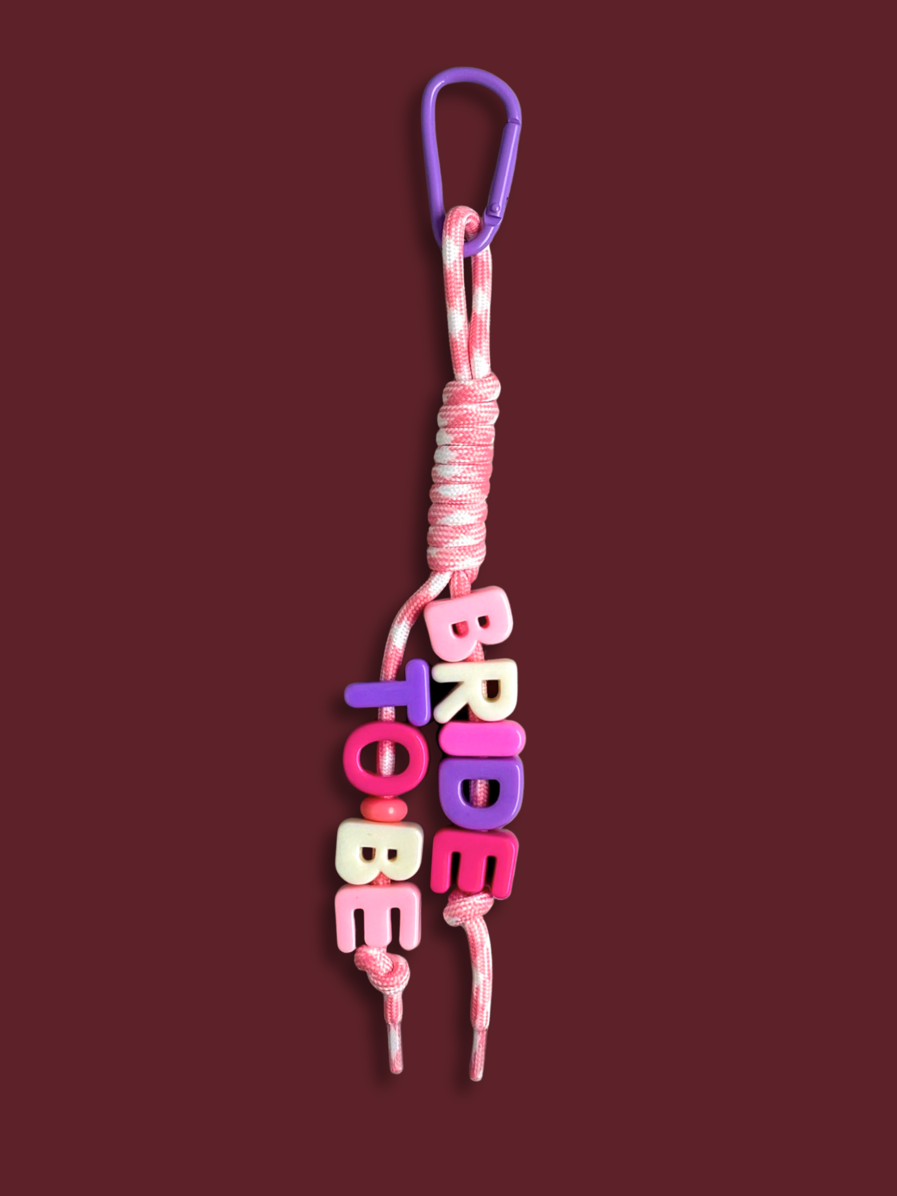 Custom statement bag charm with letter beads spelling BRIDE TO BE in pink, white and purple, threaded across two strands of a pink and white houndstooth knotted cord with a purple carabiner clip, on a burgundy background. Made by Bugalugs in Melbourne. 