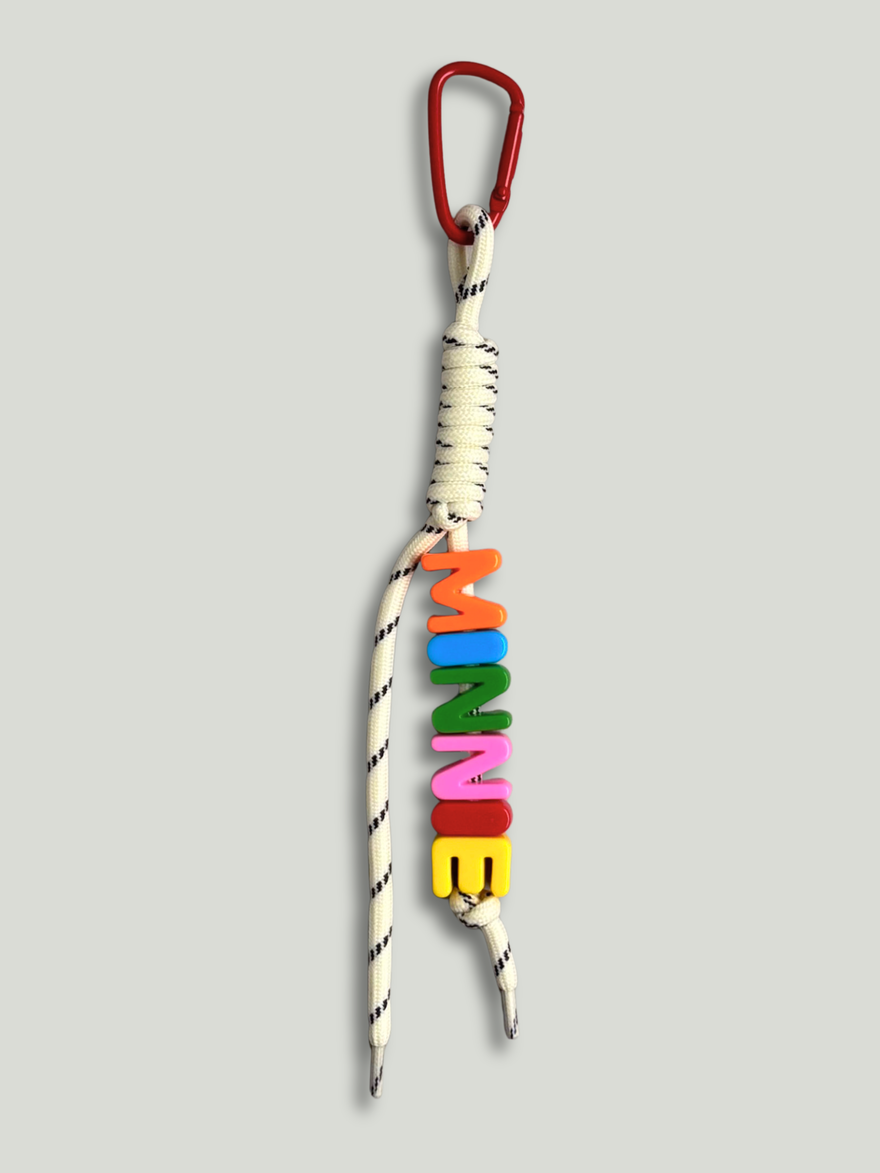 Personalised bag charm with letter beads spelling MINNIE in orange, blue, green, pink, red and yellow, threaded on a cream and black fleck knotted cord, with a red carabiner clip on a gray background. Made by Bugalugs in Melbourne.